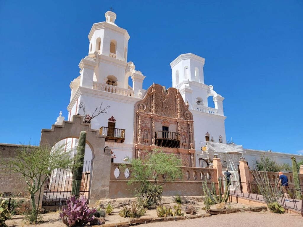 Mission San Xavier del Bac in Tucson, Arizona. Beautiful Spanish church with ornate historical architecture. The front of the building has two white towers and the center entrance area is an adobe tan color. The building is decorated with scrolled ornamentation and small sculptures. There is an arched opening in the adobe wall around the building with desert landscaping in front of it. Southwest States