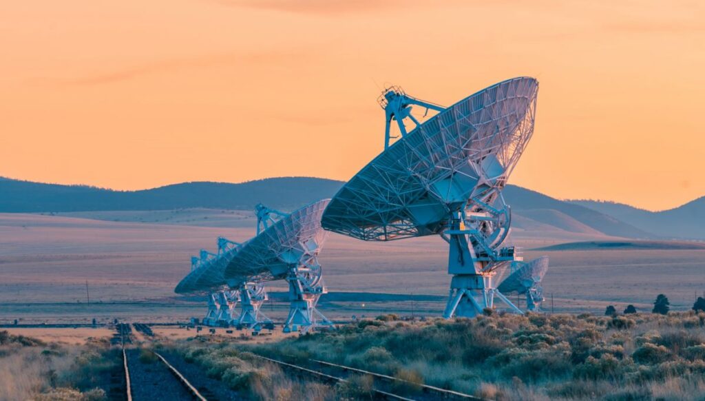 Massive radio telescopes that look like satellite dishes pointed to the sky on the remote land near Magdalena, New Mexico. Rail tracks run parallel to the 5 telescopes. The pale orange sky of the sunset is in the background. Southwest Region USA