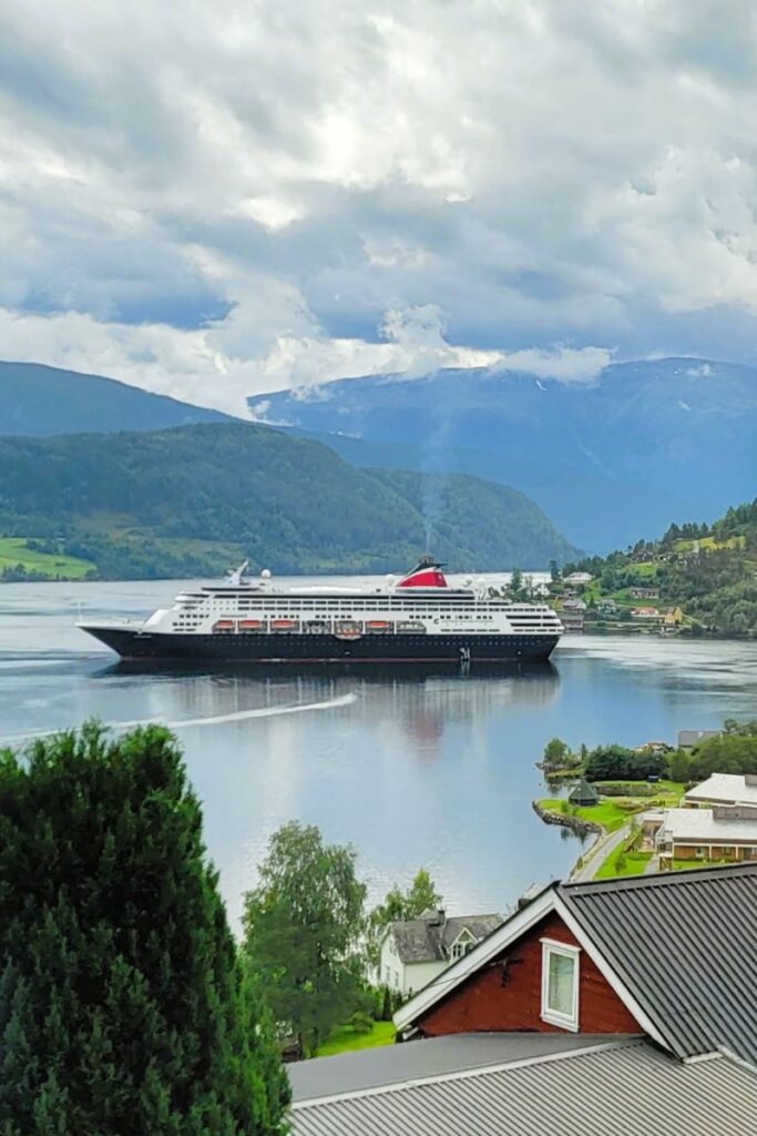 A cruise ship glides on the water at Hardanger Fjord near a small village. A red building is in the foreground. The sky is cloudy and there are rolling hills in the distance. 