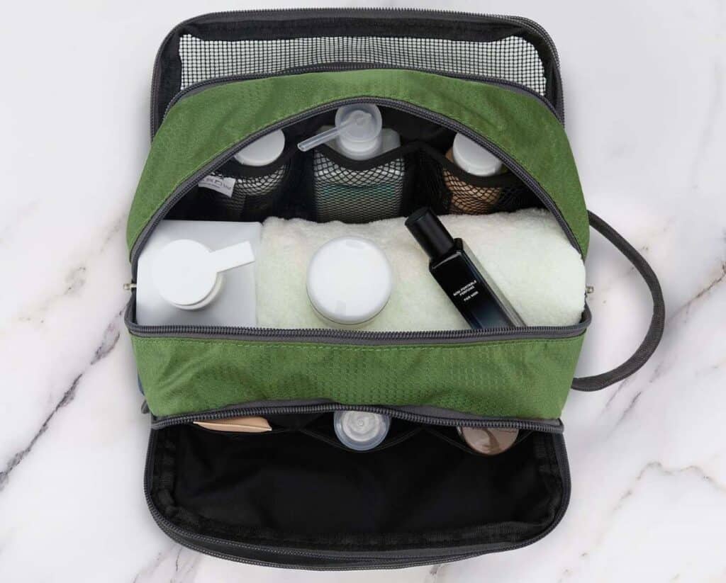 Army green toiletry travel bag with the zippers open. You can see various bottles, jars and a white washcloth inside.