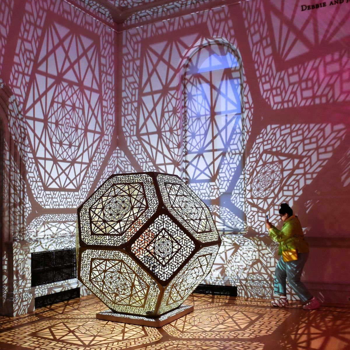 A woman takes a picture with her phone of the HYBYCOZO exhibit at the Renwick Gallery in Washington DC. The work consists of larger than life geometric sculptures, often with pattern and texture that draw on inspirations from mathematics, science, and natural phenomena. The sculptures are lit from within and reflect on walls and surrounding areas.