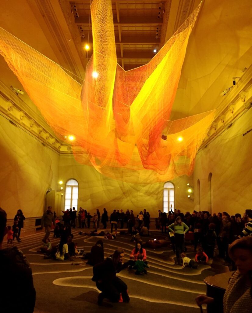 Flowing netting in colors lit up to show blends of yellow, orange and red hang from the ceiling of the Renwick Gallery. Visitors sit and lay on the floor and stand beneath the Janet Echelman's 1.8 exhibit.