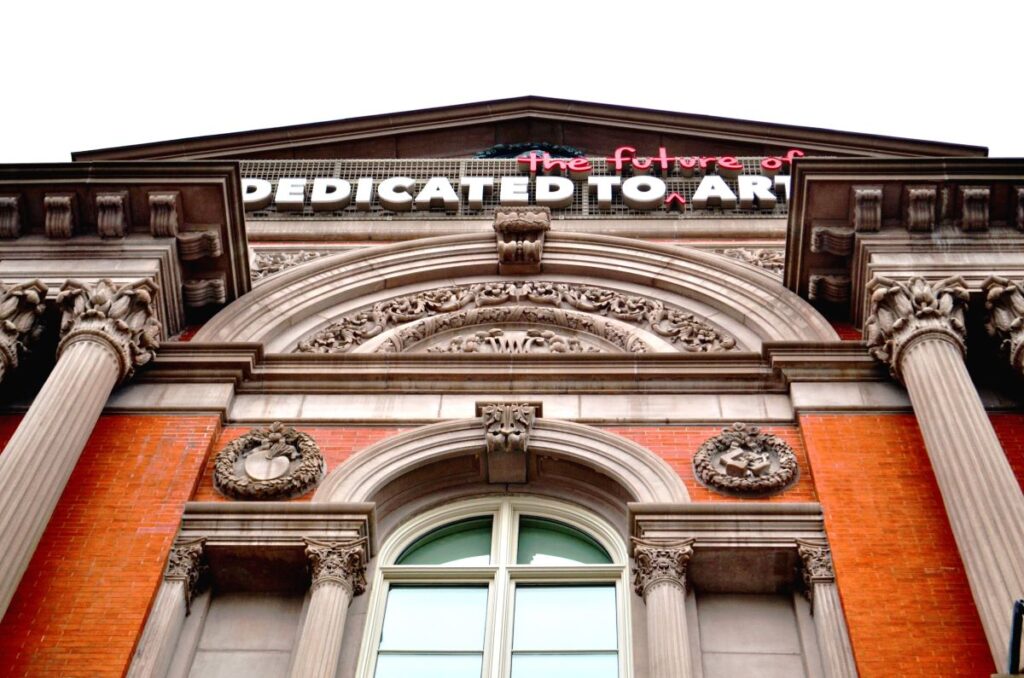 Renwick Gallery building in Washington DC. Orange brick frames an arched window with Roman-style columns and an ornate stone arch about the window. Words above say "Dedicated to the Future of Art"