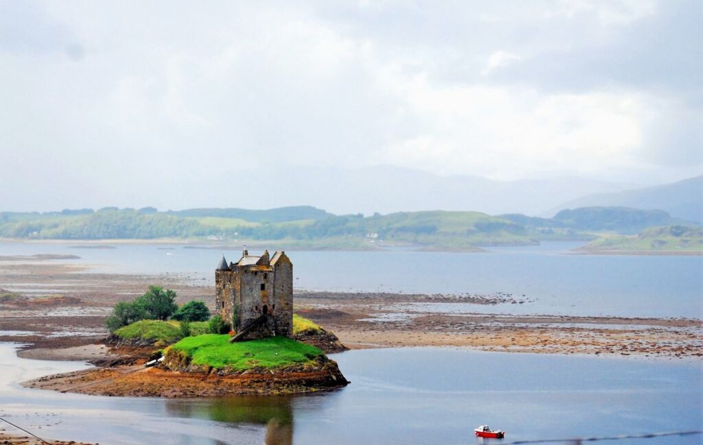 Medieval Stone castle in Scotland sits on a tiny island surrounded by a small amount of grass that is also surrounded by low water levels at low tide. A red boat moves toward the island in the water.