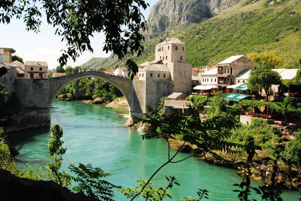 The historic Stari Most stone bridge arches over the turquoise waters of the Neretva River in Mostar, Bosnia and Herzegovina, framed by lush greenery and medieval architecture, making it a meaningful travel destination.