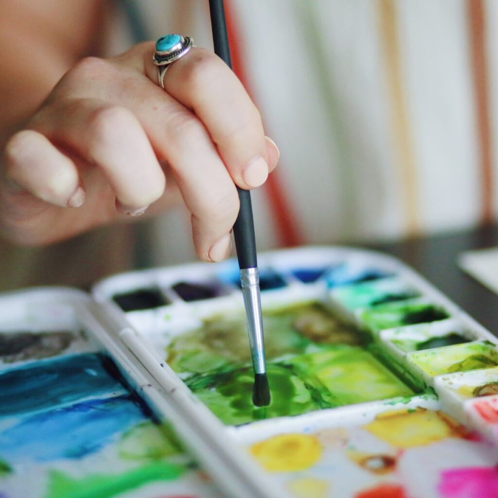 Woman's hand holding a paint brush, mixing watercolor paint in a tray. 