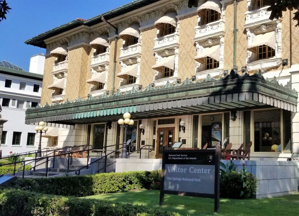 A tan 3-story building with a covered front veranda. There is a tan canvas awning over each window. The sign in front of the building says Hot Springs National Park Visitor Center Arkansas. Great stop for an American road trip