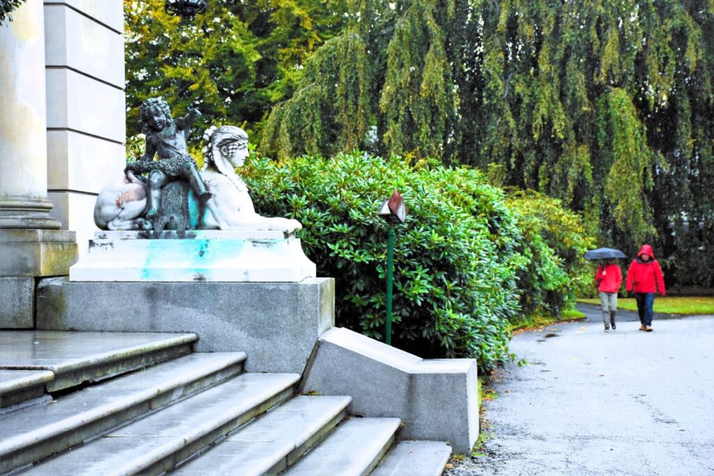 Two people in red jackets, one with an umbrella, walk toward the marble steps of The Elms mansion in the rain. A marble sculpture of a cherub sitting on a lion like creature is next to the steps.