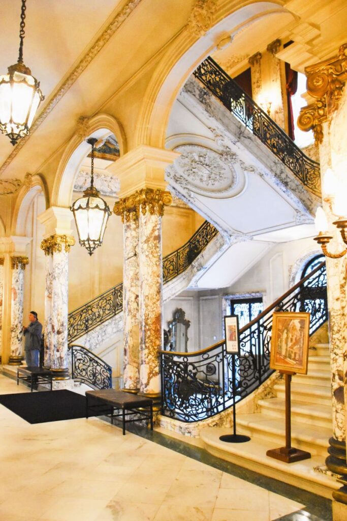 Spectacular grand double staircase with ornate black wrought iron curved railings and marble steps. A man stands near one of the six marble columns that can be seen on either side of the staircase. The Elms Gilded Age mansion in Newport RI USA