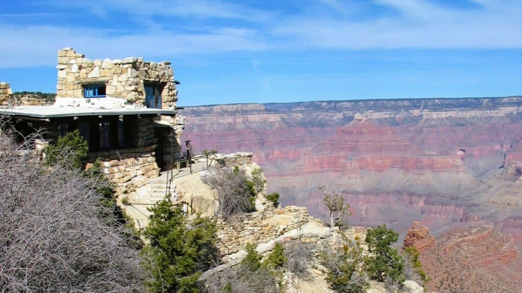 "A rustic stone building with blue-trimmed windows sits on the edge of the Grand Canyon's South Rim. The structure is built from local limestone that blends with the rocky cliff face. The vast canyon stretches in the background, showing red and orange layered rock formations under a blue sky with wispy clouds."
