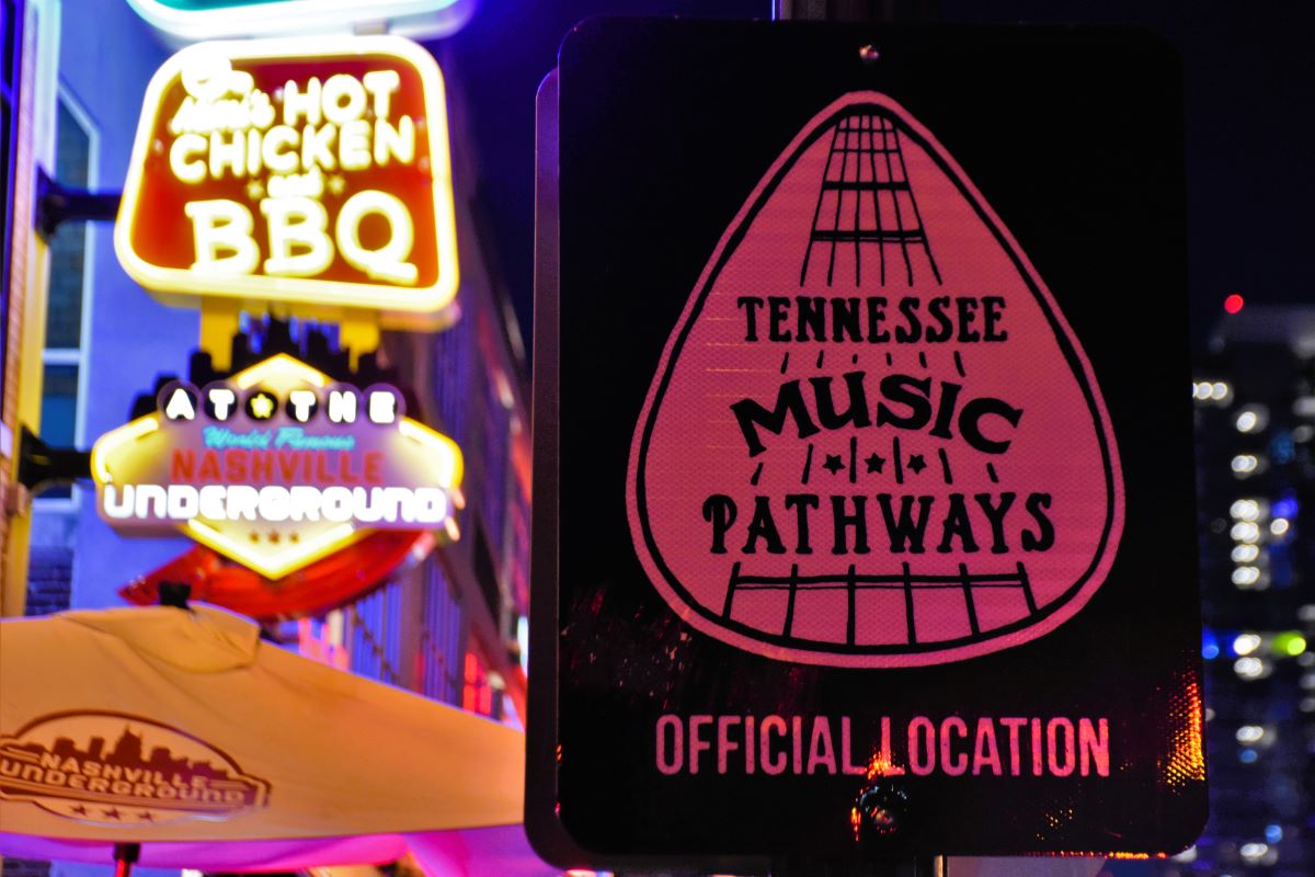 배경에는 여러 개의 밝은 네온 불빛과 BBQ를 포함한 다양한 간판, Tennessee Music Pathways 공식 위치라고 적힌 간판이 있습니다.