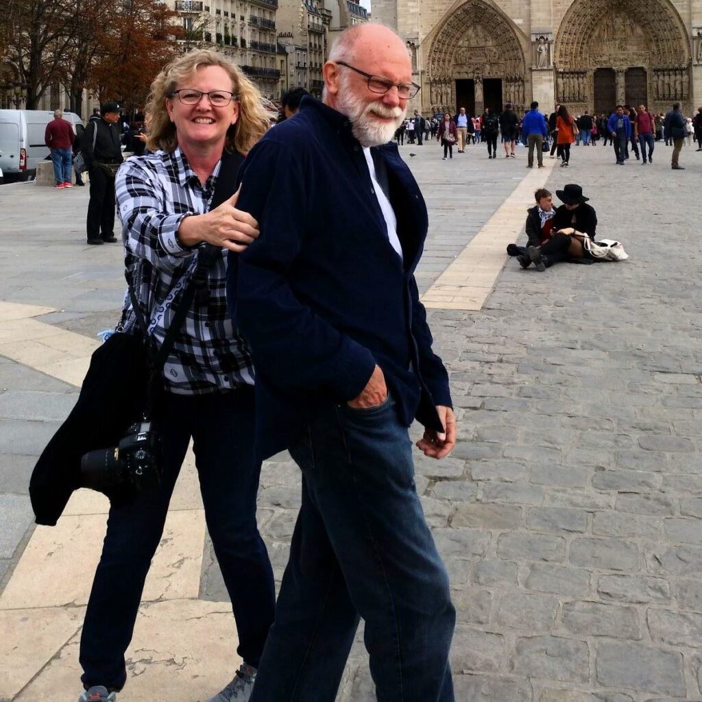 Blonde woman is laughing while she grabs the arm of her husband (a man with a white beard) to try to get him to pose for a picture in front of the Notre Dame Cathedral in Paris France.