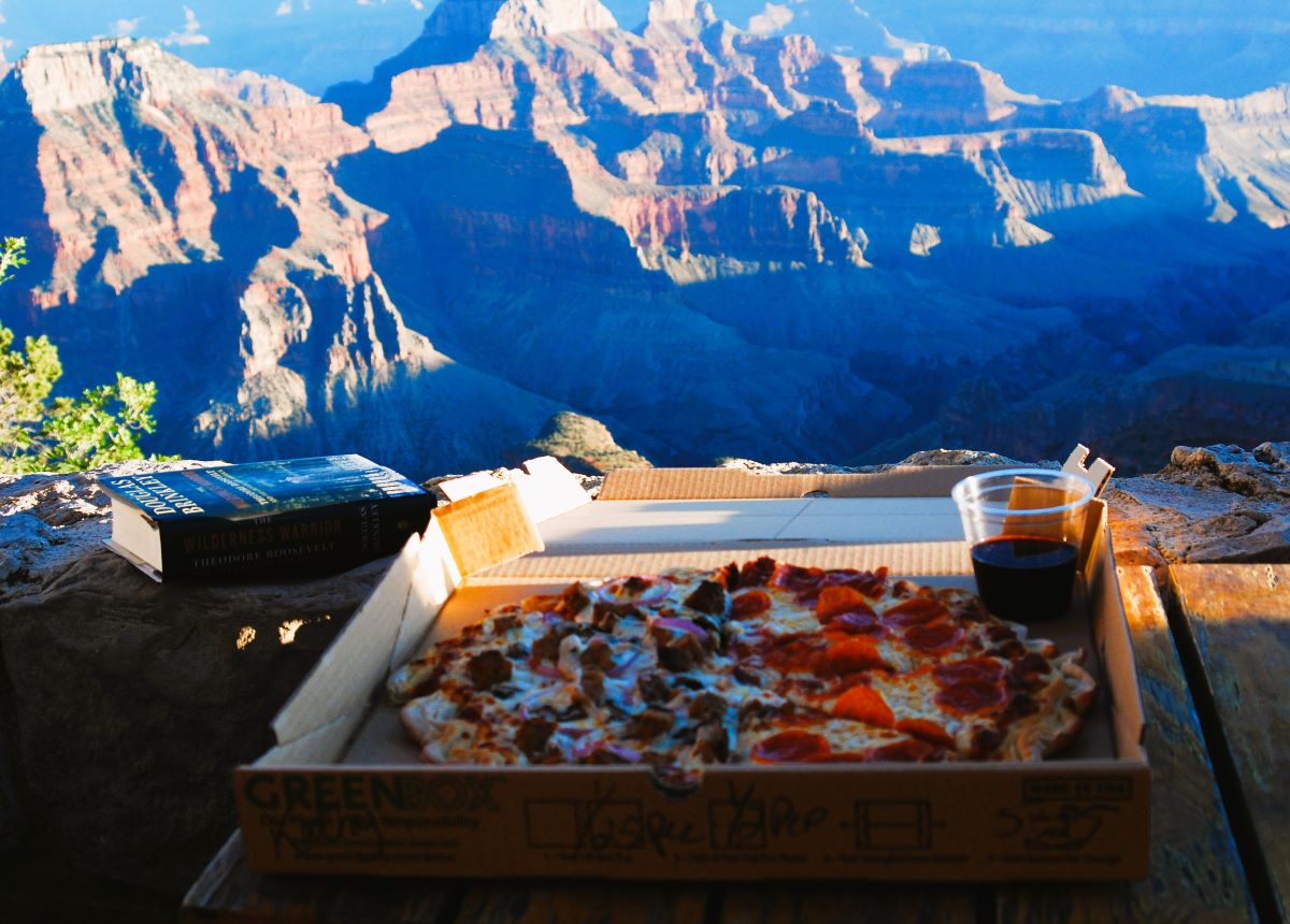 A pizza with half pepperoni and half sausage sits in a pizza box on a rustic wood table. A clear plastic cup holds red wine and sits in a corner of the pizza box. A thick hardcover book "The Wilderness Warrior" sits on a stone wall next to the pizza box and the Grand Canyon at sunset is in the background.