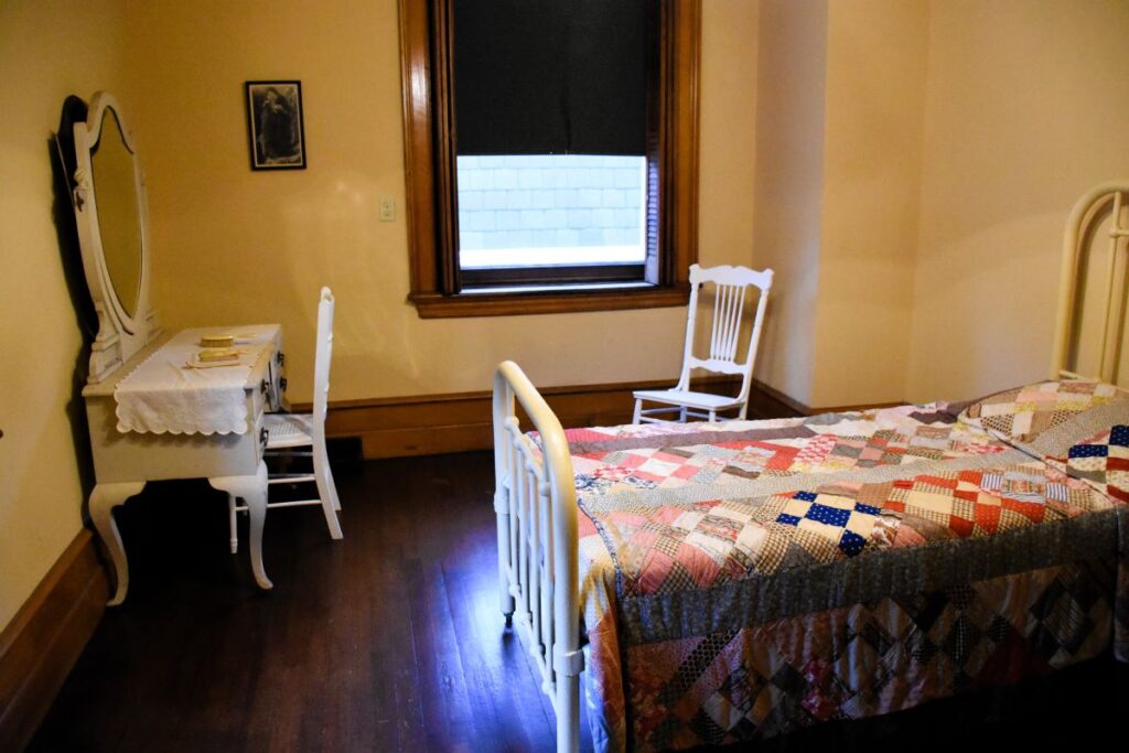 Servant's quarters at the Elms Mansion - A simple white iron bed with a patchwork quilt on top. A simple white vanity with mirror and a wooden chair sits across the room. The floor is wood with no rug. Newport, Rhode Island