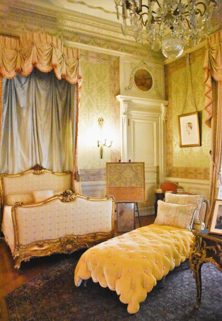 Newport Mansions - an opulent yellow bedroom with an ornate canopy over the bed and velvet covered chaise lounge in front. This room is at the Elms Mansion in Newport Rhode Island. Mrs. Berwind’s Bedroom appears as the room of the Russells’ daughter, Gladys in the TV show, The Gilded Age.