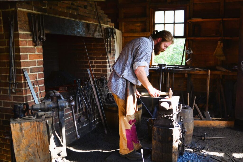 A blacksmith working in a traditional workshop at Mount Vernon, George Washington's estate in one of the Southeast states in the USA. The blacksmith hammers a heated metal piece on an anvil next to a brick fireplace forge, with various tools hanging on the wall and sunlight streaming through the window.