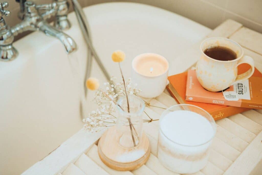 A small wooden tray sits on top of a clawfoot bathtub. The tray holds a lit candle, a cup of coffee in a patterned mug, a small vase with dried flowers, and two round objects that could be coasters or lids. The vase and candle are placed on wooden discs, and an open book titled ‘Daily Rituals’ is visible. The setting suggests a cozy and tranquil environment, perfect for reading or enjoying a warm beverage. Self care is perfect for when you're bored.