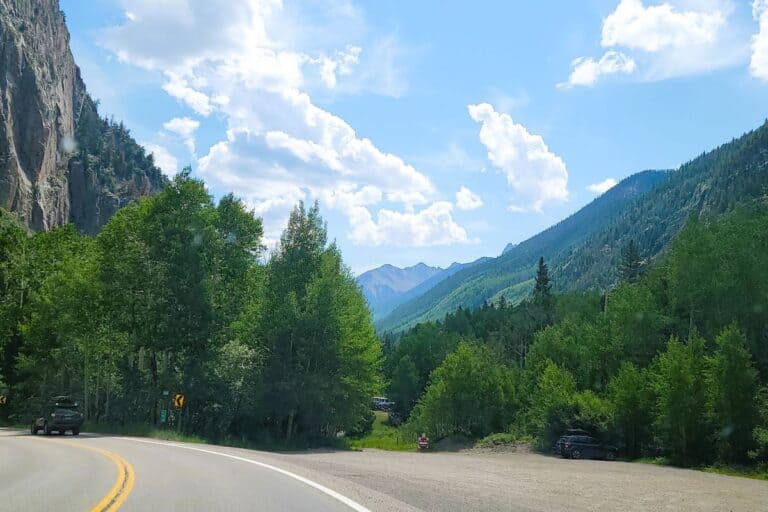 A scenic view of a car driving on a winding mountain road surrounded by lush green trees and steep cliffs under a bright blue sky with fluffy white clouds. The road curves gently to the left, with vehicles parked along the roadside and distant mountain peaks visible in the background, creating a serene and picturesque landscape.