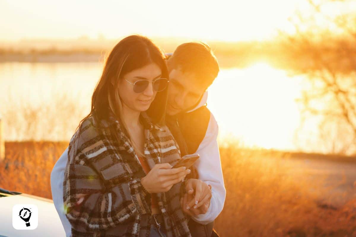 A couple enjoying a sunset while looking at their smartphone. The background shows a glowing sun with shades of gold, orange and brown reflecting on a lake, evoking a peaceful road trip moment.