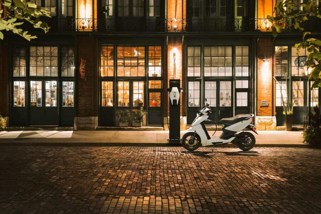 A white electric scooter sits charging at an EV charging station on a cobblestone street, in front of a historic building with large glass windows warmly lit from inside at night.