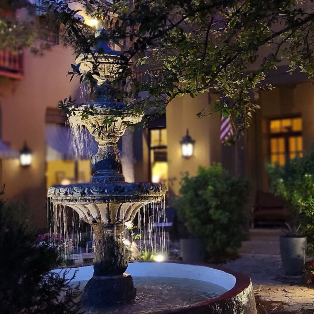 A 4-tier fountain flows at twilight in the middle of the courtyard at Hotel El Capitan in Van Horn, Texas. A tree branch, American flag and windows can be seen in the background.