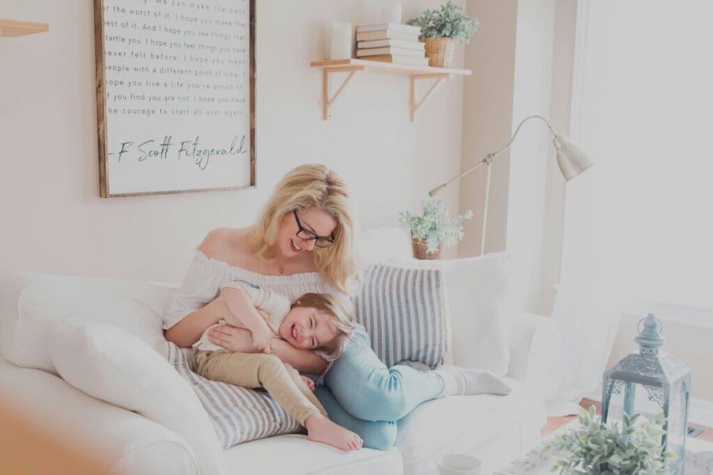 A bright and cozy living room scene with a smiling woman wearing glasses, sitting on a white sofa, playfully interacting with a young child who is lying across her lap. The room has a soft color palette with light walls, and there’s decorative text on the wall behind them. A wooden shelf with minimal decor items is visible in the background.