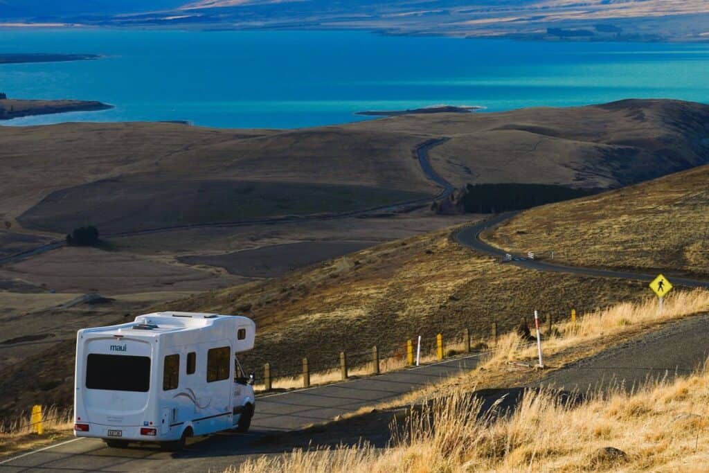 A white RV driving down a winding road through dry, rolling hills with a view of a bright blue lake in the distance under a clear sky. A pedestrian crossing sign is visible along the roadside.