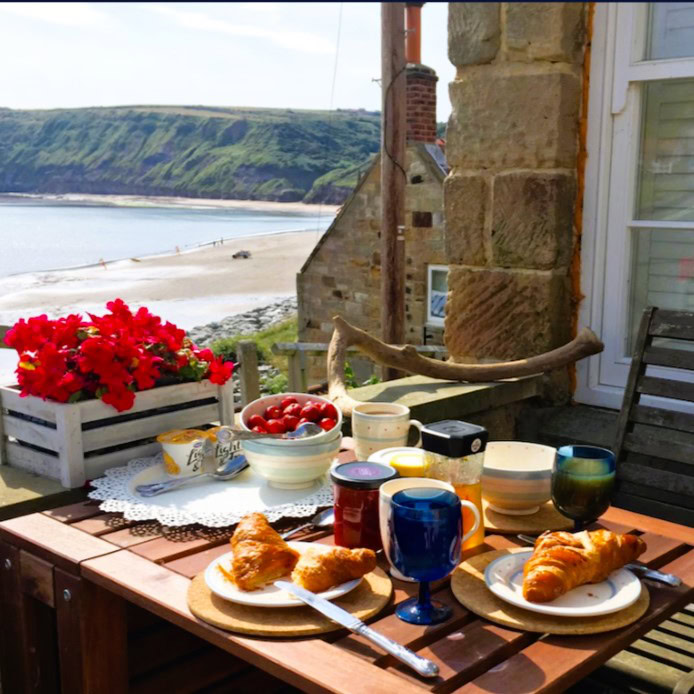 A stone cottage with an outdoor patio and wooden table. Breakfast croissants, strawberries and beverage glasses sit on the table. A pot of red flowers sits on the stone wall next to the table with a view of the ocean. VRBO