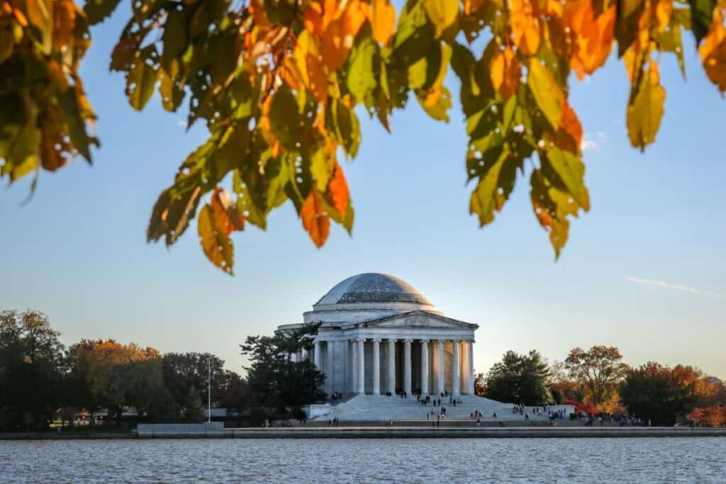 The Jefferson Memorial in Washington, D.C., is seen from across the Tidal Basin. The round structure features a domed roof and columns, with people visible on the steps and around the monument. The foreground includes autumn leaves in shades of green, yellow, and orange, framing the top of the image.