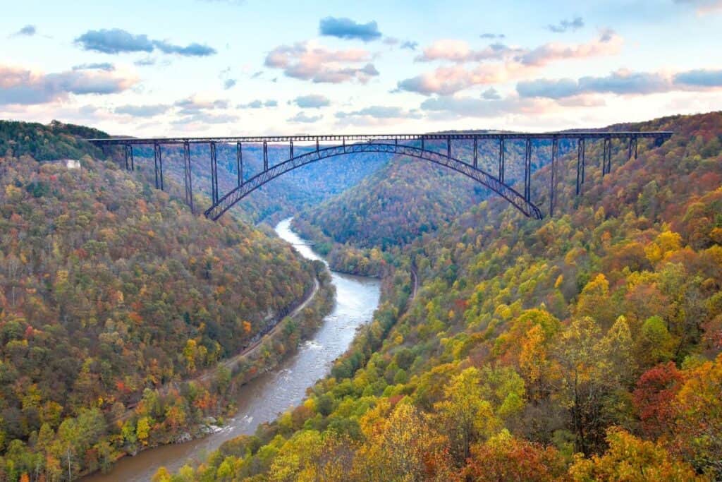 New River Gorge Bridgeis one of the best national parks in autumn. Spanning across the vibrant fall foliage of New River Gorge National Park, West Virginia. The steel arch bridge stands tall against a backdrop of colorful trees in shades of green, orange, yellow, and red, with the New River flowing below under a partly cloudy sky.