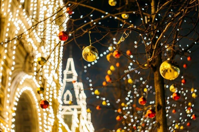 Close-up of festive holiday lights and ornaments on tree branches, with golden baubles reflecting the glow of string lights in a cityscape at night, evoking seasonal holiday warmth and cheer.