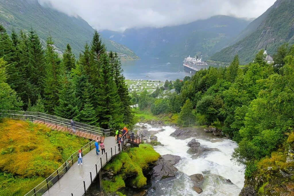 People stand on a trail next to a waterfall on a cloudy day. The small town of Geiranger Norway is seen in the distance and a Holland America cruise ship floats in the port with steep mountains on all sides.