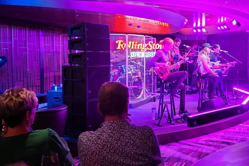 Two adults sit in a cruise ship nightclub with their back to the camera watchin a live band of 5 young adults playing guitars and various instruments. The stage is lit in purplish lighting.