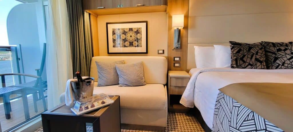 A queen size bed, a bottle of champagne sits on a small table in front of a loveseat with artwork hanging behind it and a deck chair on the balcony of a Holland America Cruise Line Balcony Suite on Norway cruise - get cruise tips for choosing the best cabin