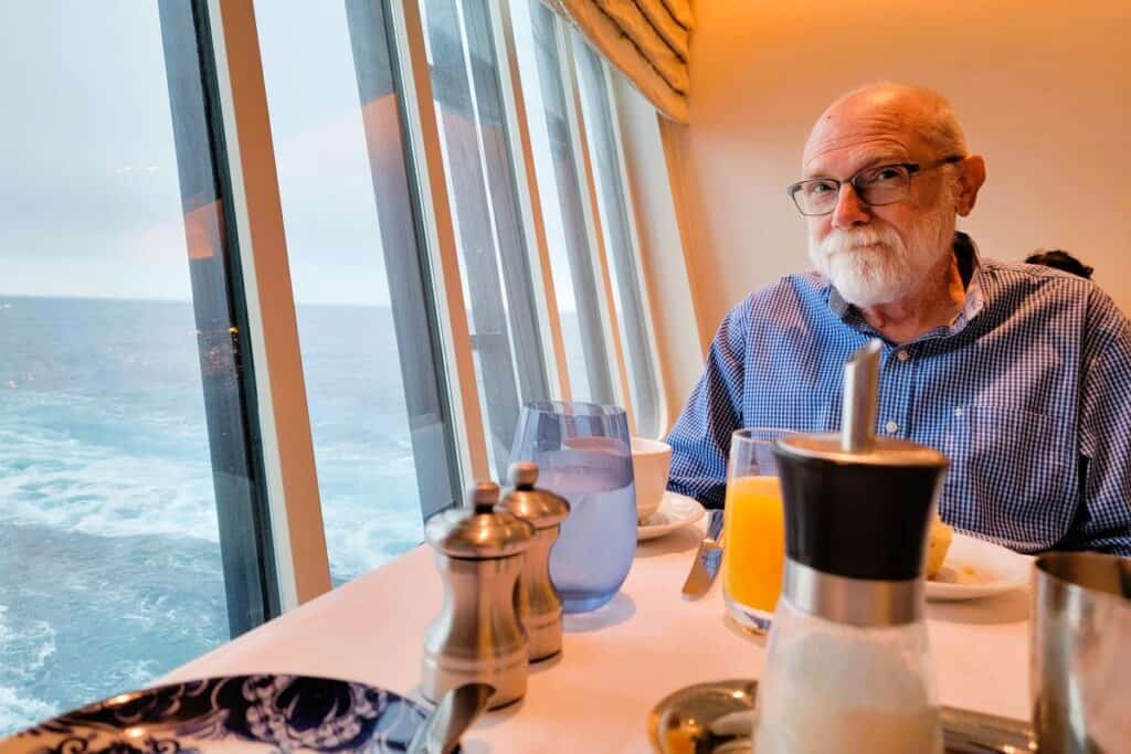 Older man with a white beard sits at a restaurant table with a white tablecloth and a glass of orange juice. He is dining on a cruise ship and the ocean waves can be seen out the window next to the table.