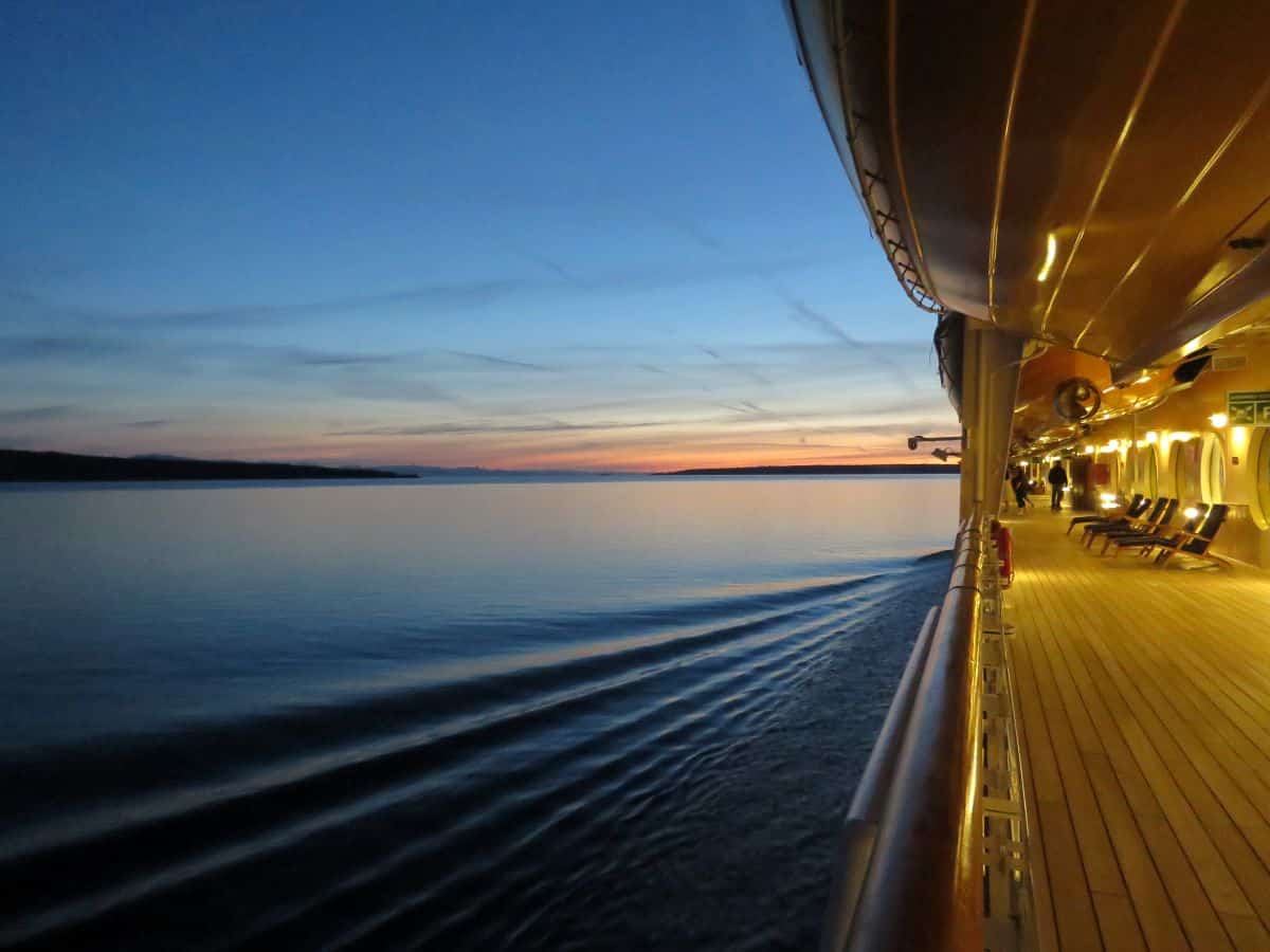 View from standing on the deck of a cruise ship. Smooth water and a clear sky at sunset can be seen. Also a quiet wooden deck with empty deck chairs and a few people walking in the distance.